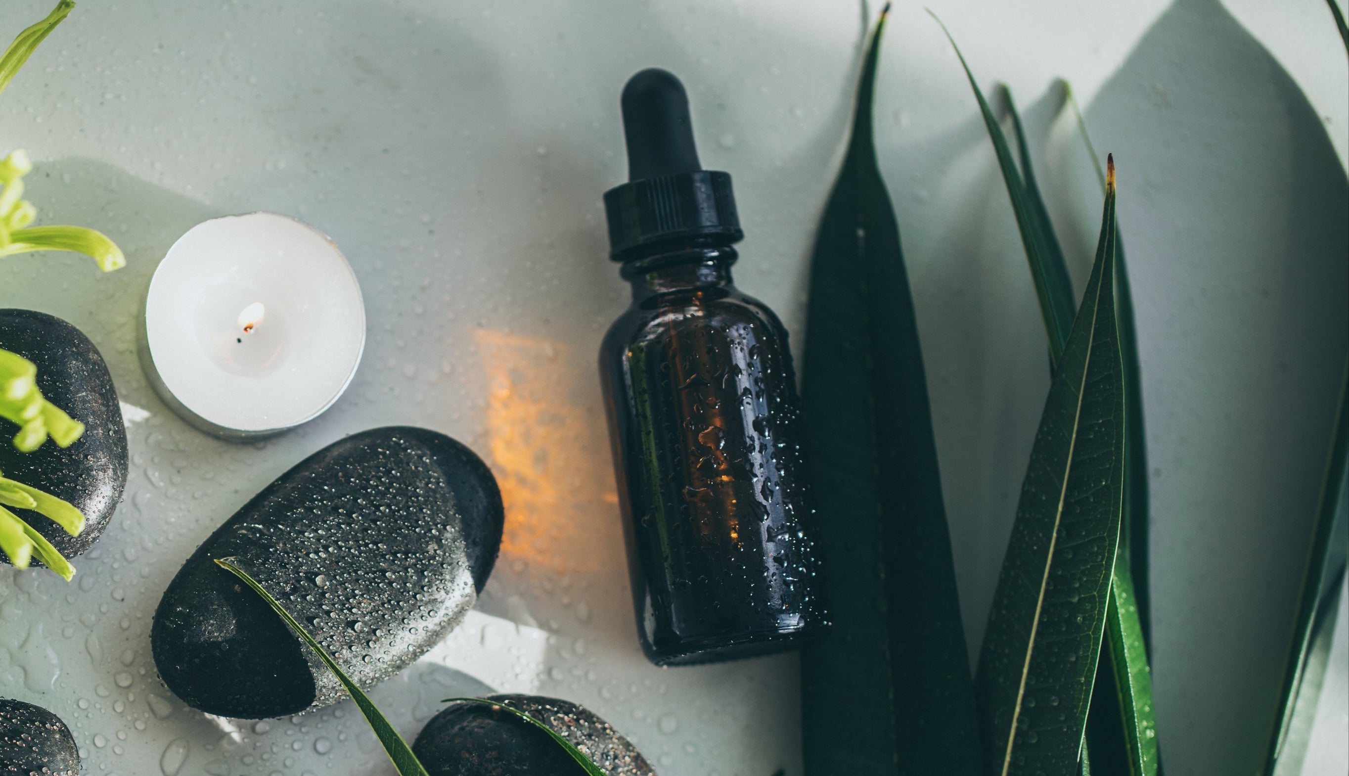 Spa setting with stones, candles, a plant, and a dropper bottle on a white surface.
